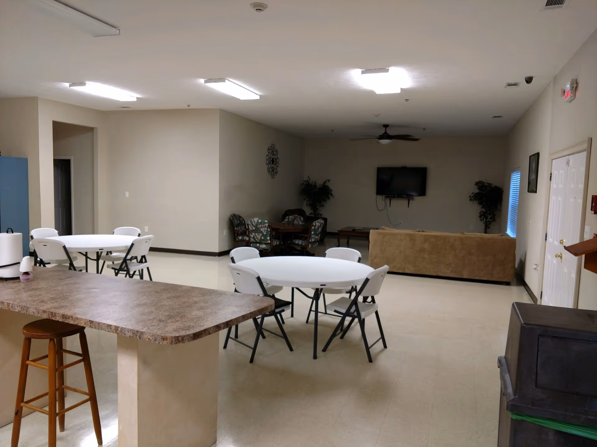 A spacious room with round tables and folding chairs arranged for seating, a countertop with a wooden stool, a beige couch facing a wall-mounted TV, and some potted plants. The room has a ceiling fan and fluorescent lighting.