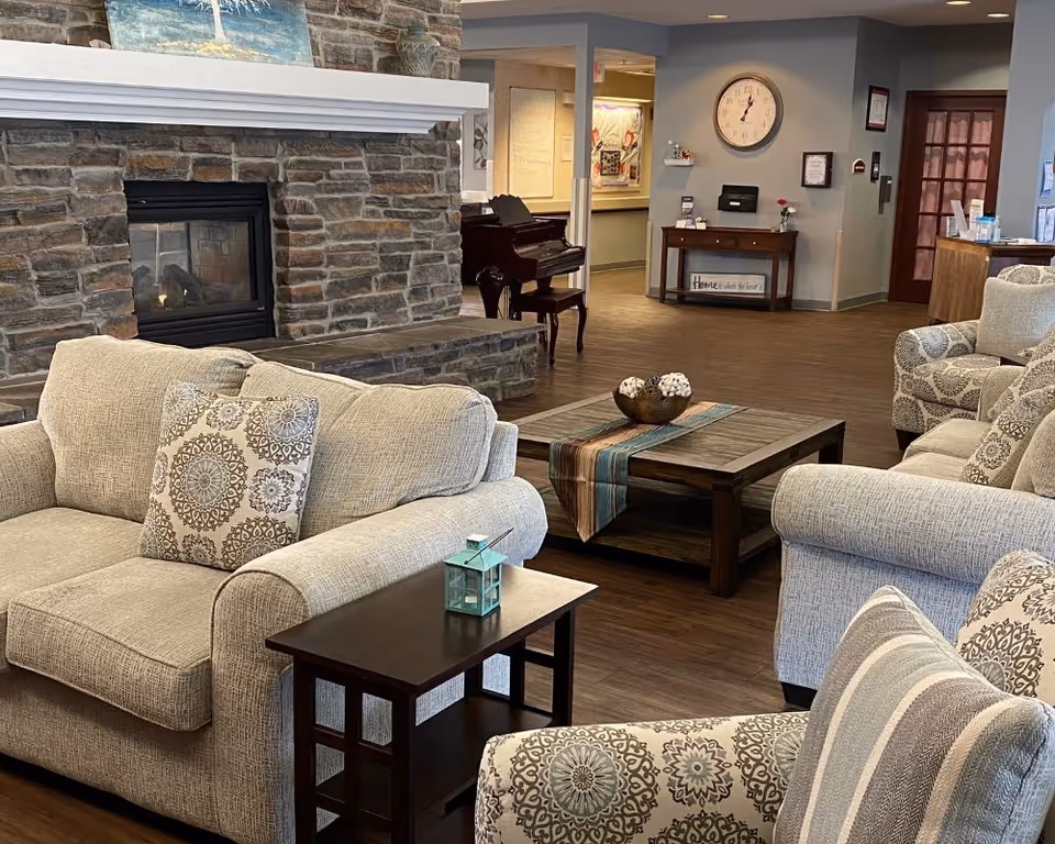 A cozy living room area with beige upholstered sofas and armchairs featuring patterned cushions. A wooden coffee table with a decorative runner and bowl sits in the center. Behind the seating area is a stone fireplace with a white mantle, and a piano is visible in the background. The room has wooden flooring and neutral-colored walls with a clock and framed items on the far wall.