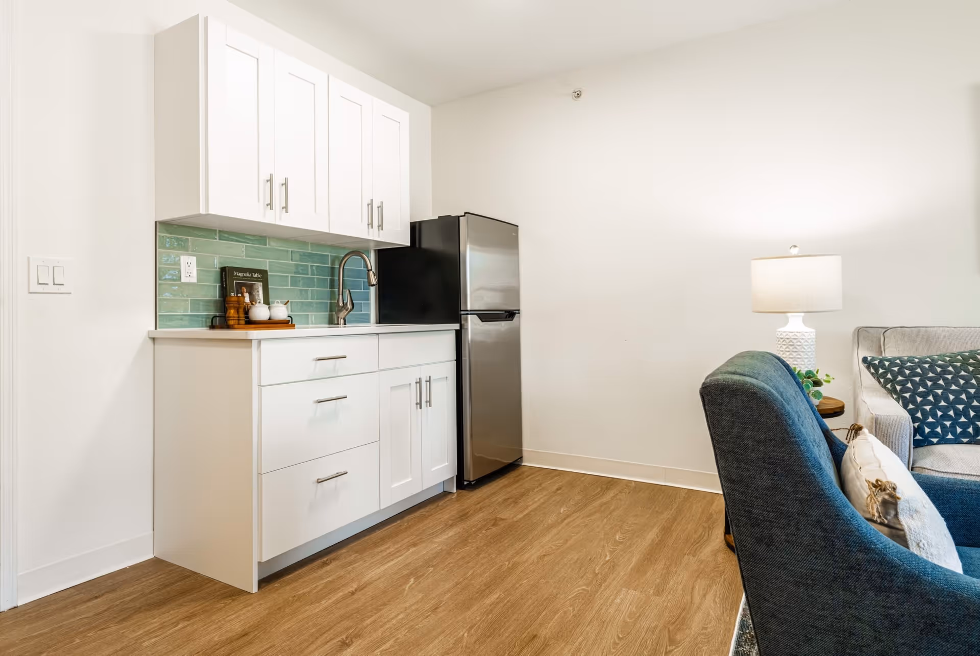 A small kitchen area with white cabinets, a stainless steel refrigerator, a sink with a modern faucet, and a green tiled backsplash. Adjacent to the kitchen is a living area with a blue upholstered chair, a side table with a white lamp, and part of a gray sofa with patterned cushions.