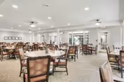 A spacious dining room with multiple round tables and cushioned chairs arranged neatly. The room is well-lit with ceiling lights and natural light coming through large windows and glass doors leading outside. The walls are light-colored, and the carpeted floor adds a cozy feel to the space.