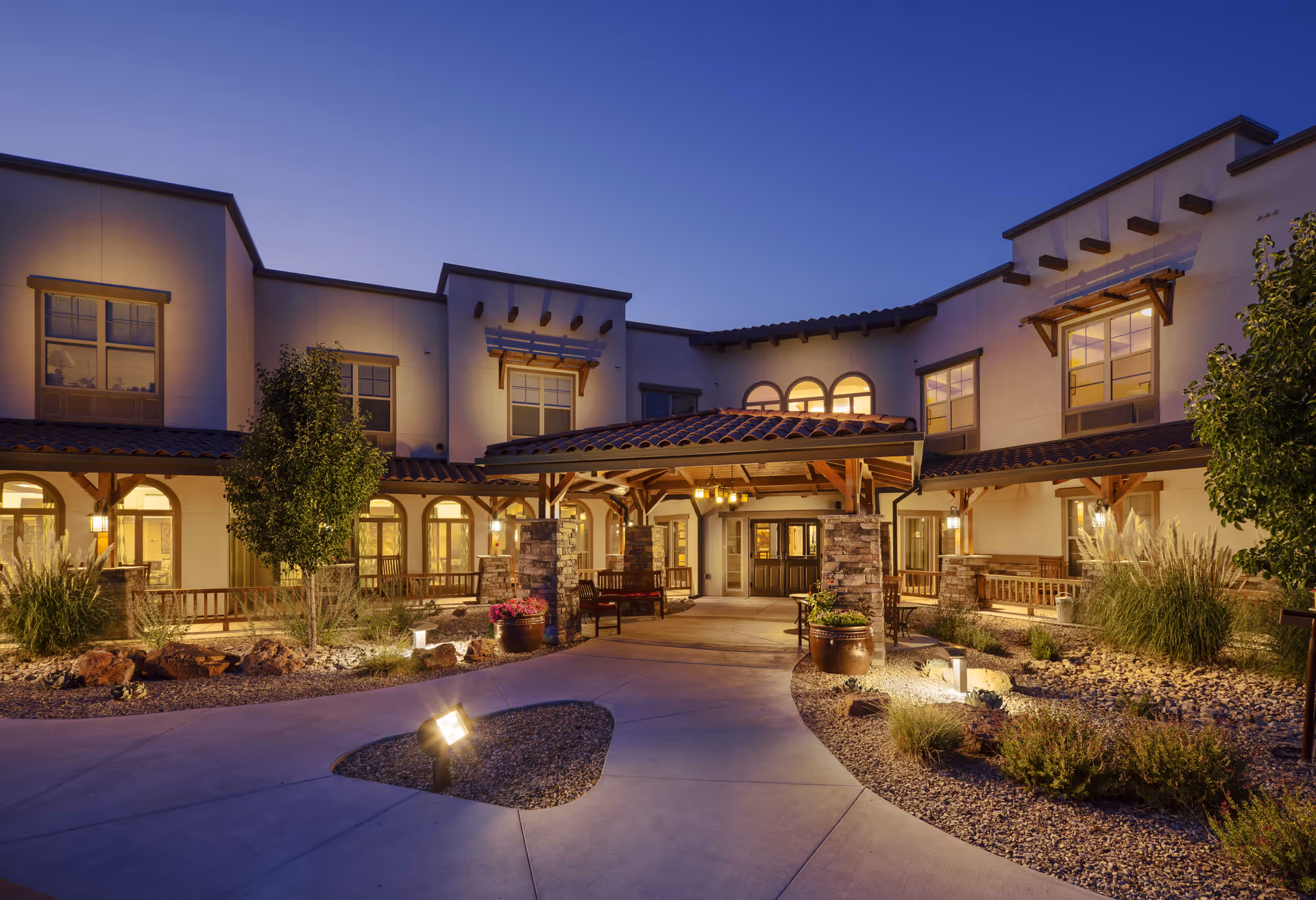 Dusk view of a lit assisted-living building front with a covered entryway, pathways and landscaped courtyard.