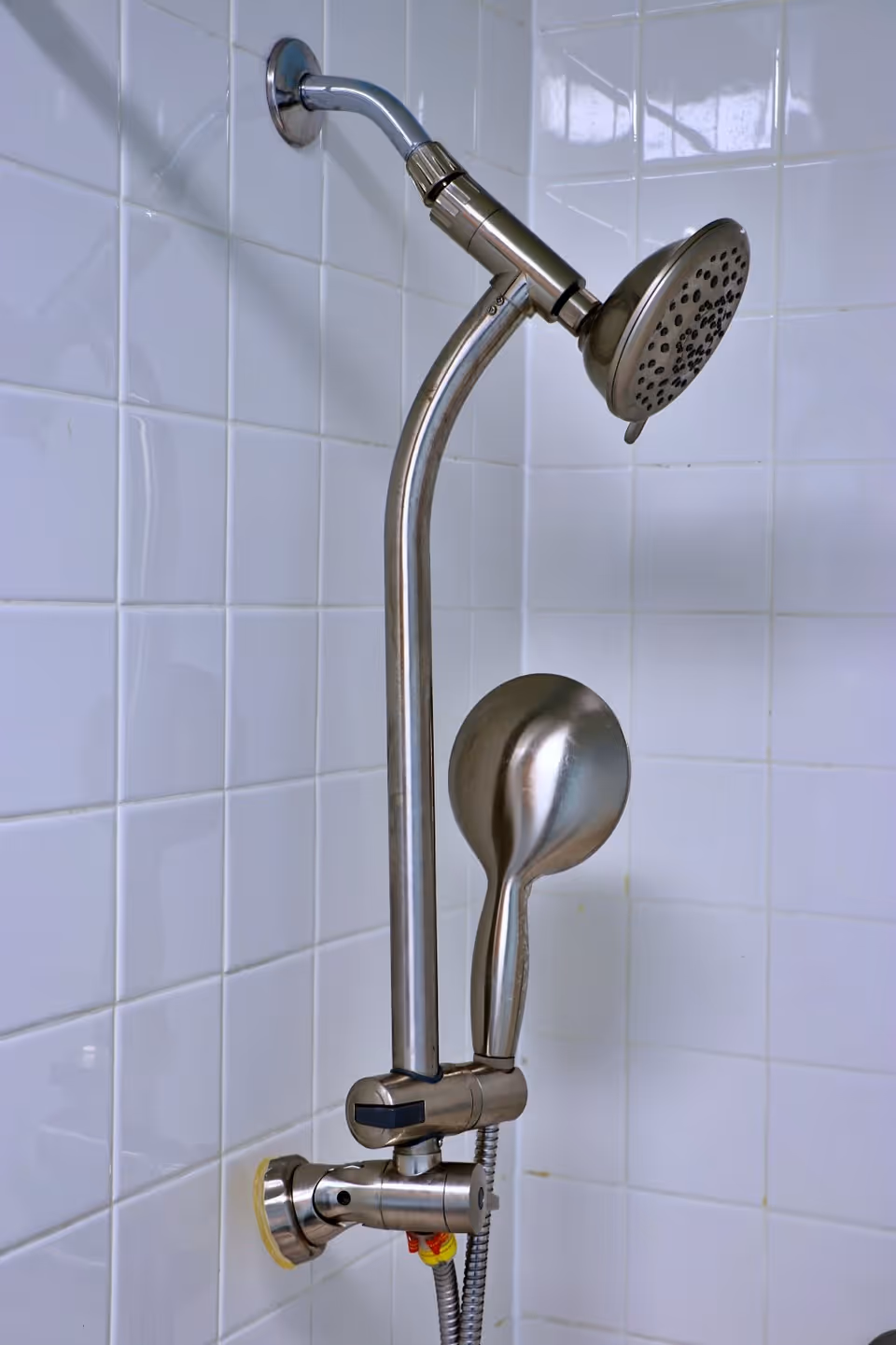 Close-up view of a modern stainless steel shower fixture mounted on a white tiled wall, featuring a fixed showerhead and a detachable handheld showerhead.