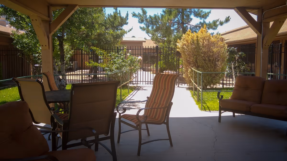 Covered outdoor patio area with cushioned chairs and a table, overlooking a gated garden with trees and shrubs under a clear blue sky.