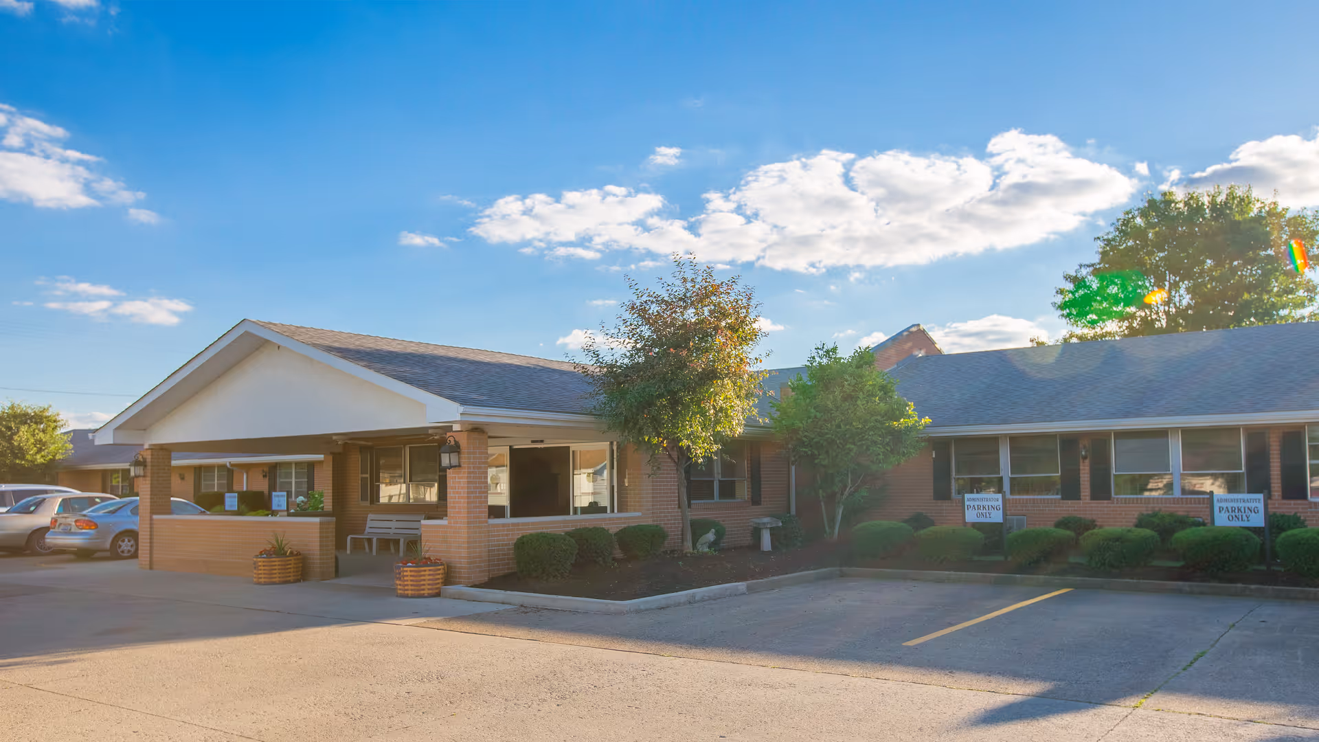 Exterior view of Arlington Care Center building with a covered entrance, brick walls, several windows, and a parking area with cars. There are small trees and bushes in front of the building under a partly cloudy blue sky.