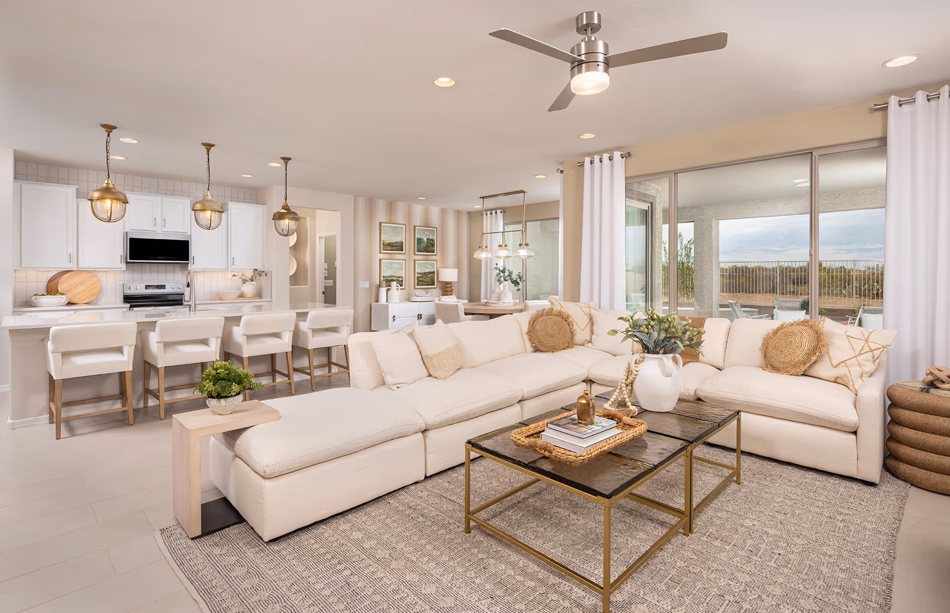 Bright and spacious open-concept living room and kitchen area with a large white sectional sofa adorned with decorative pillows, a glass coffee table with books and a vase, and a kitchen island with four white bar stools. The kitchen features white cabinetry, stainless steel appliances, and three pendant lights. Large sliding glass doors lead to an outdoor patio with a scenic view.