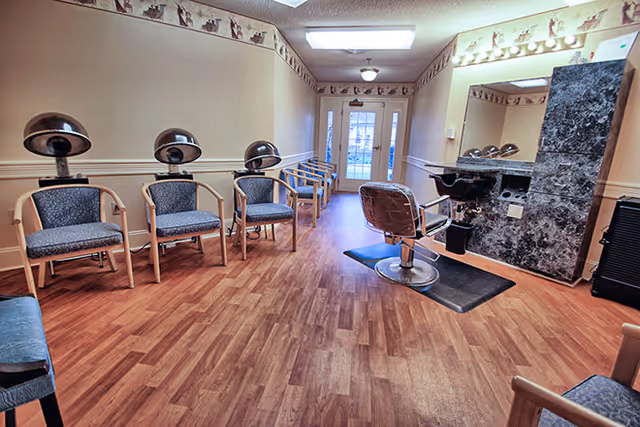 Interior view of a salon area in a senior living facility with multiple hair drying chairs lined up against the left wall, a single styling chair in front of a large mirror and sink on the right, wood flooring, and a door with glass panels at the far end.