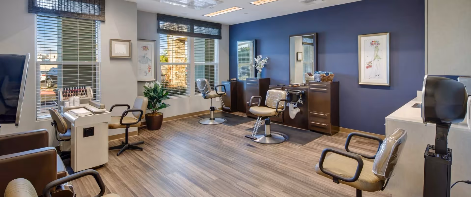 Interior of a salon area with multiple styling chairs, mirrors, and manicure stations. The room has large windows with blinds, a blue accent wall, wooden flooring, and framed artwork on the walls.