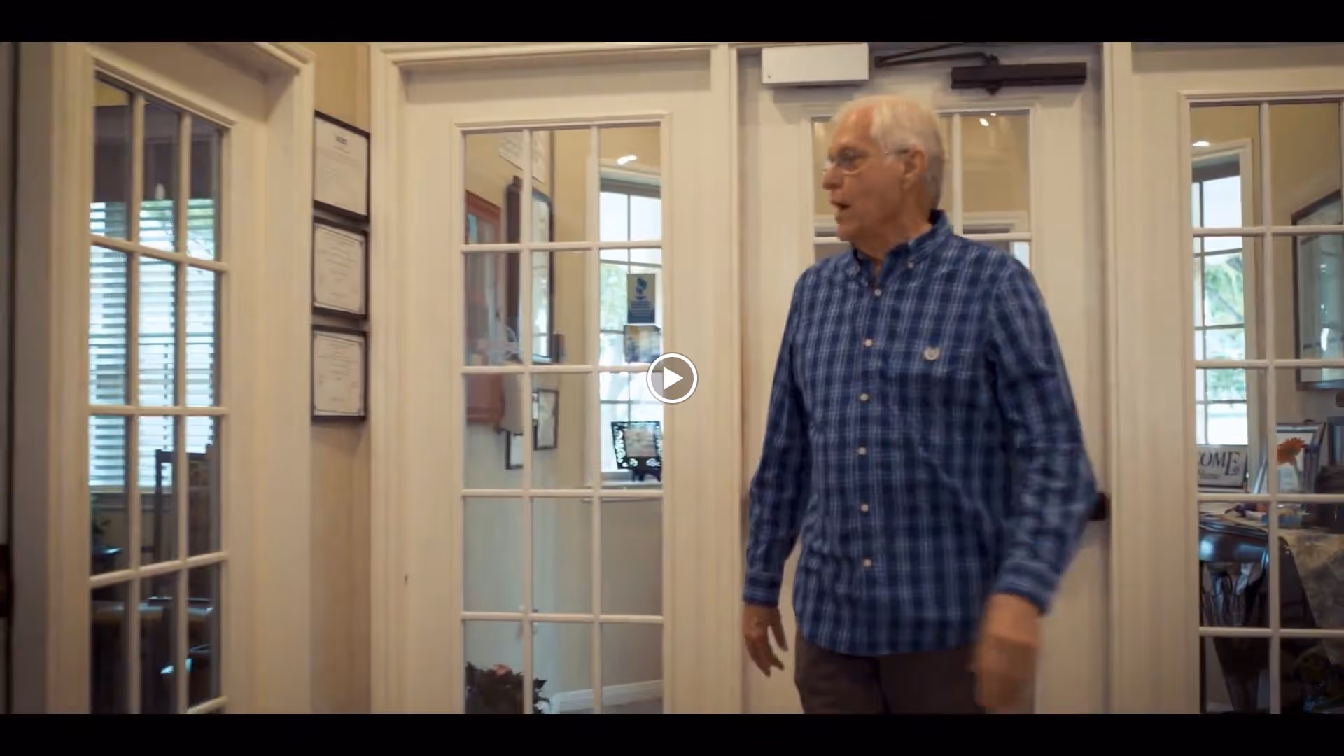An elderly man wearing a blue plaid shirt stands in a hallway with glass-paneled doors on either side. The hallway has cream-colored walls with framed certificates or documents hanging on the left wall. Through the glass doors, parts of a room with windows and furniture are visible.