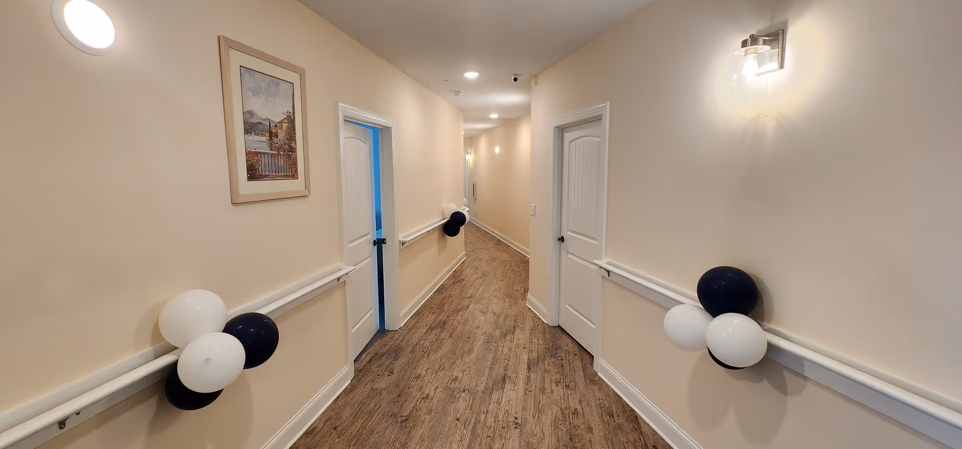 A well-lit hallway in a senior care facility with light beige walls, wooden flooring, and white doors on both sides. The hallway is decorated with clusters of black and white balloons attached to the handrails along the walls. A framed picture hangs on the left wall, and ceiling lights illuminate the corridor.