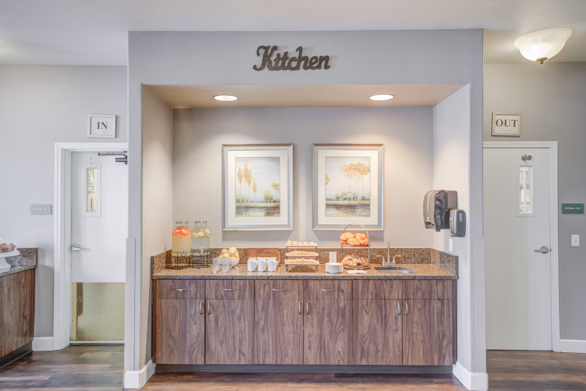 A kitchen area with wooden cabinets and granite countertop, featuring two framed landscape paintings on the wall. The countertop holds glass dispensers with beverages, cups, plates, and trays of pastries. Above the area is a sign that reads 'Kitchen'. There are two doors on either side labeled 'IN' and 'OUT'.