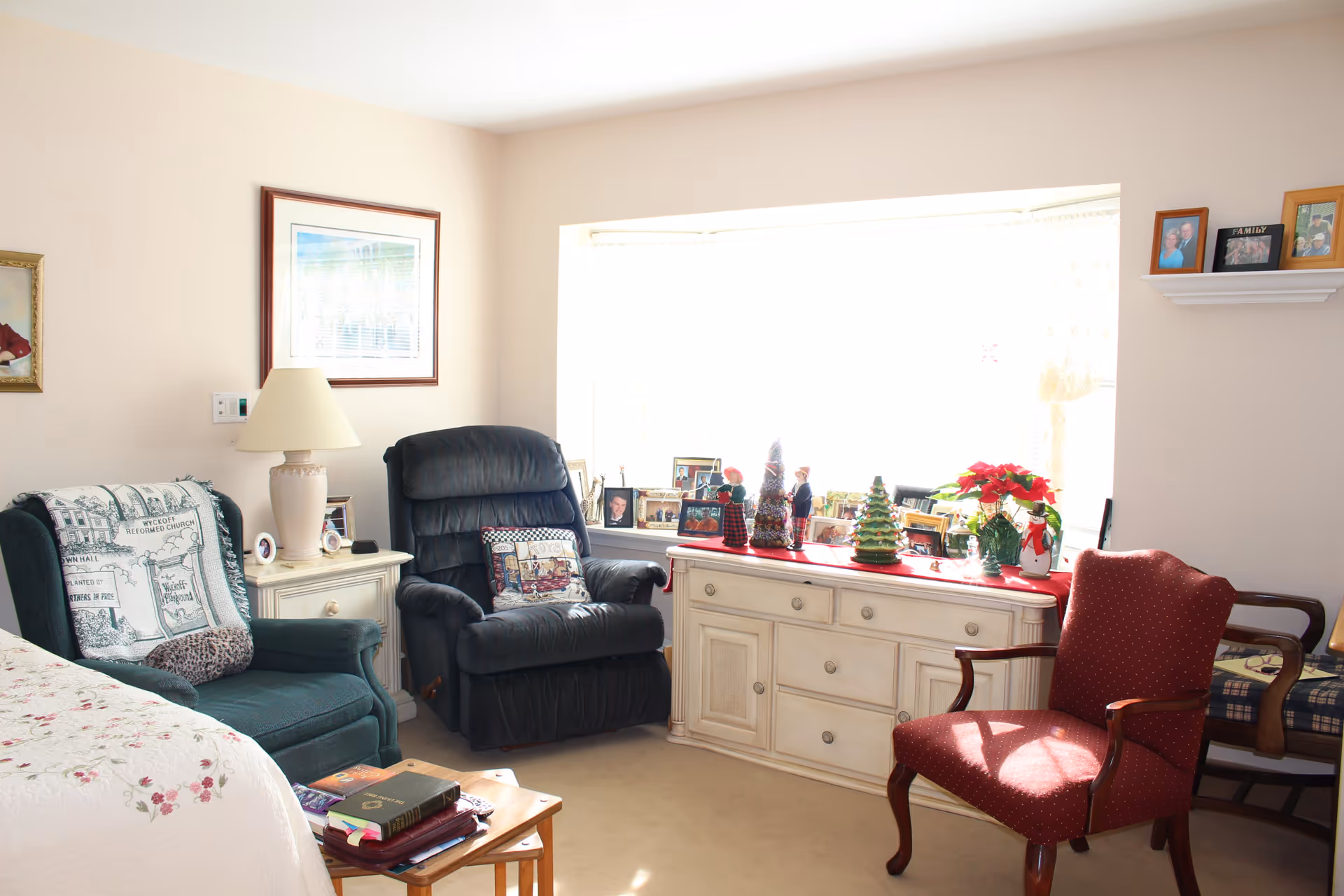 A cozy living room area with a large window letting in bright sunlight. The room features a dark green armchair with a patterned throw, a dark recliner with a decorative pillow, a red upholstered armchair, and a wooden side table with books on it. A cream-colored cabinet under the window is decorated with holiday figurines and framed photos. Family photos are also displayed on a small wall shelf.