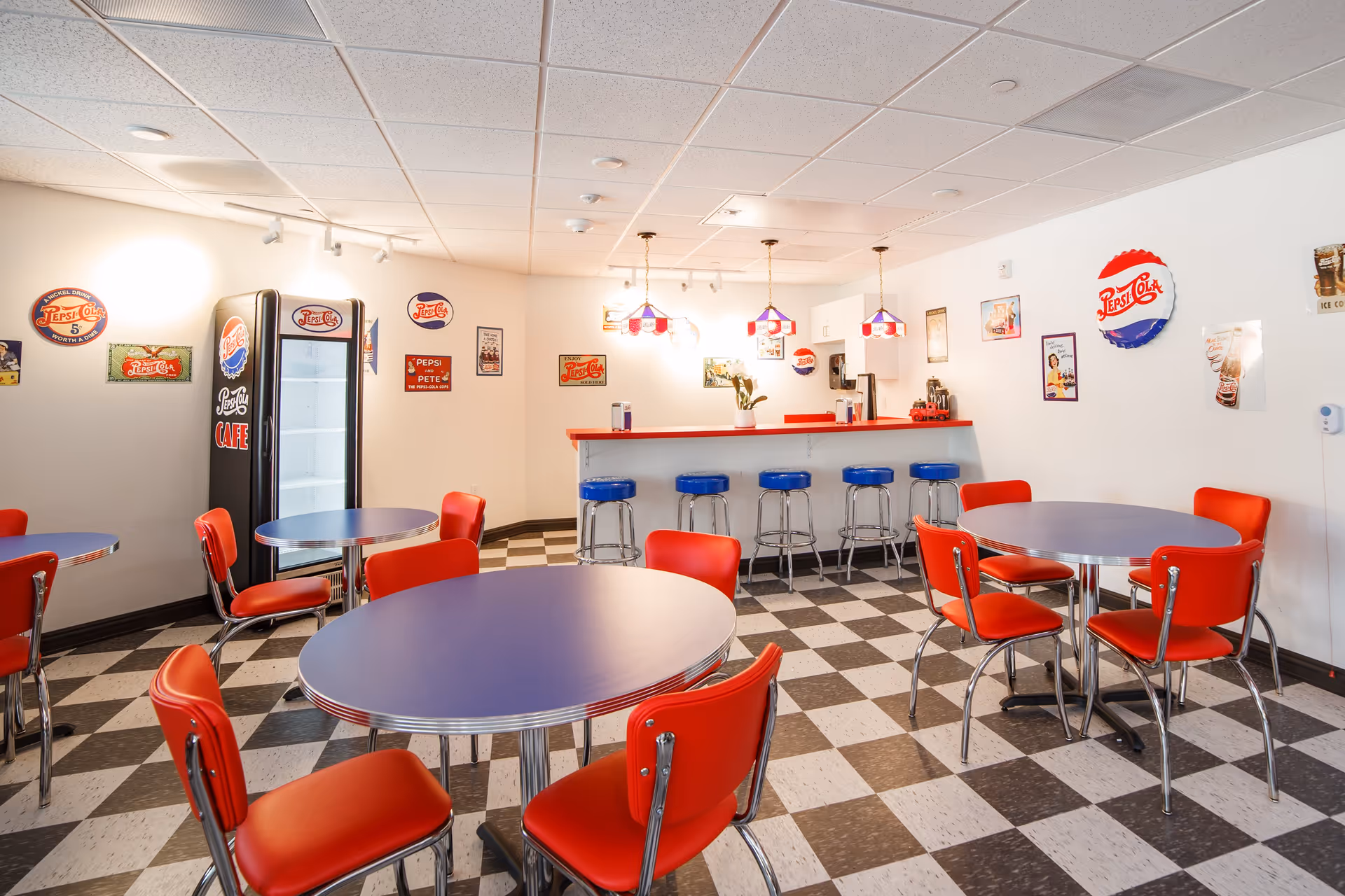 A retro-style dining area with round blue tables and red chairs on a black and white checkered floor. The room features a counter with blue bar stools and red countertop, decorated with vintage Pepsi-Cola signs and memorabilia on the walls. Pendant lights with red, white, and blue shades hang above the counter.