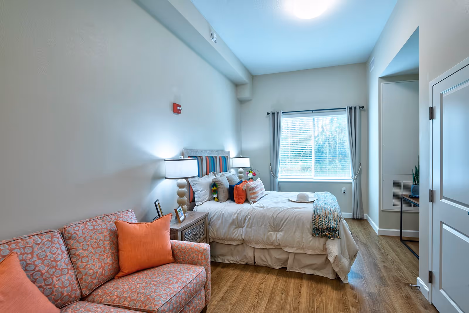 A bright bedroom with a bed featuring multiple pillows and a throw blanket, a bedside table with a lamp and picture frames, an orange patterned couch with orange pillows, a window with curtains, and wooden flooring.