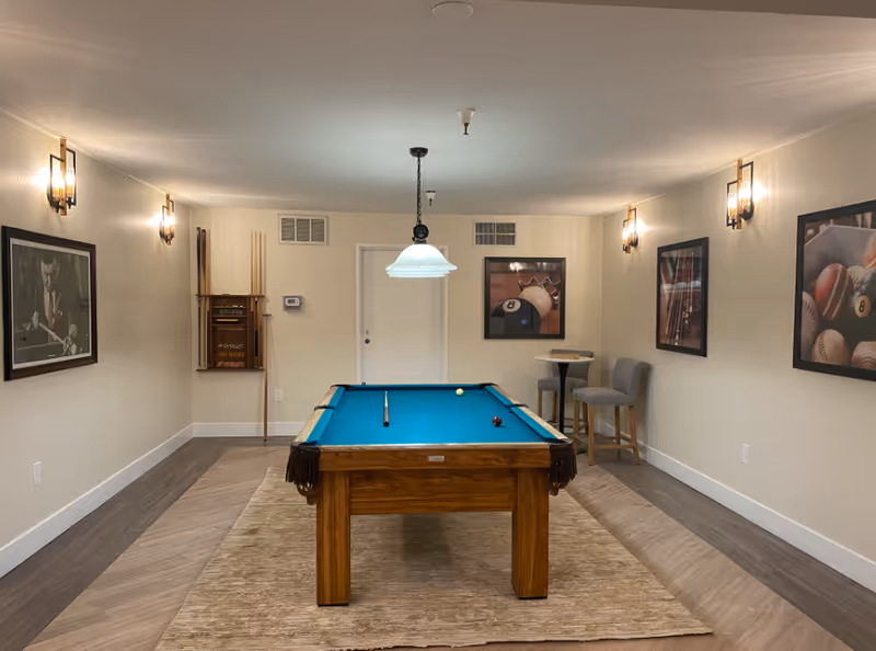 A recreation room with a pool table centered under a hanging light, framed sports artwork on the walls, and seating in the back.