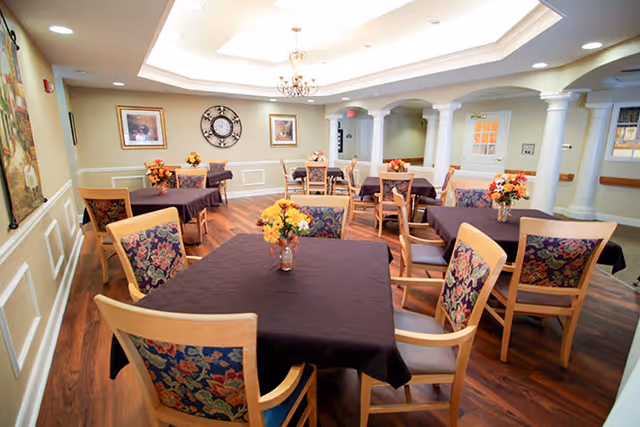 A dining room with multiple square tables covered with dark tablecloths, each table decorated with a vase of yellow and orange flowers. The room features wooden chairs with floral upholstery, a wooden floor, cream-colored walls with white wainscoting, a large decorative clock on the far wall, framed artwork, and a ceiling with recessed lighting and a chandelier.