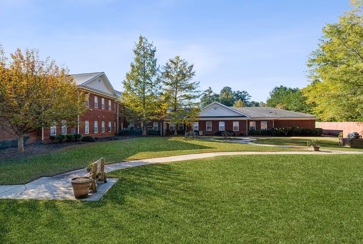 Outdoor view of a senior living facility named Northstar Square featuring a large green lawn, a curved concrete pathway, a wooden bench with a potted plant, several trees with autumn foliage, and a two-story red brick building with multiple windows under a clear blue sky.