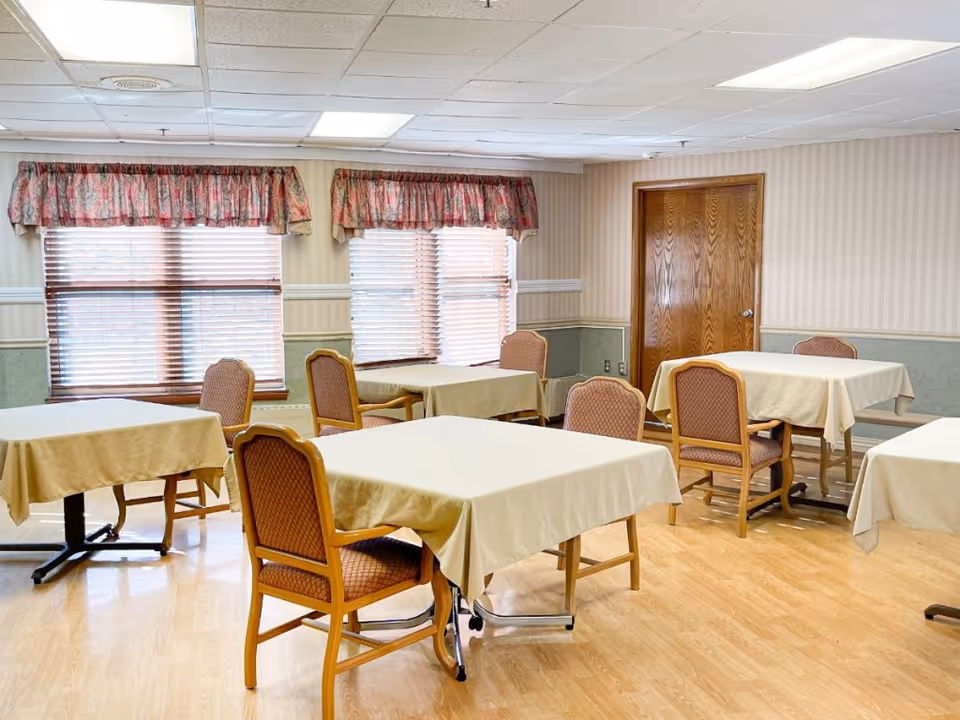 Bright communal dining room with several square tables covered in beige tablecloths and upholstered chairs near windows with blinds and floral valances.