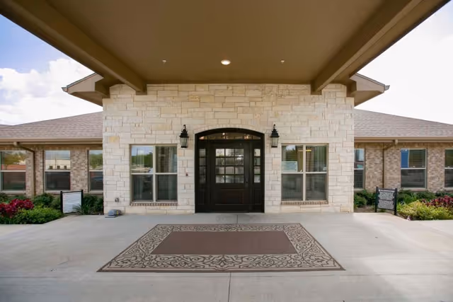 Entrance of a healthcare and rehabilitation facility with a covered driveway, stone facade, black double doors, two wall-mounted lantern lights, windows on either side, and a decorative outdoor rug on the concrete ground.