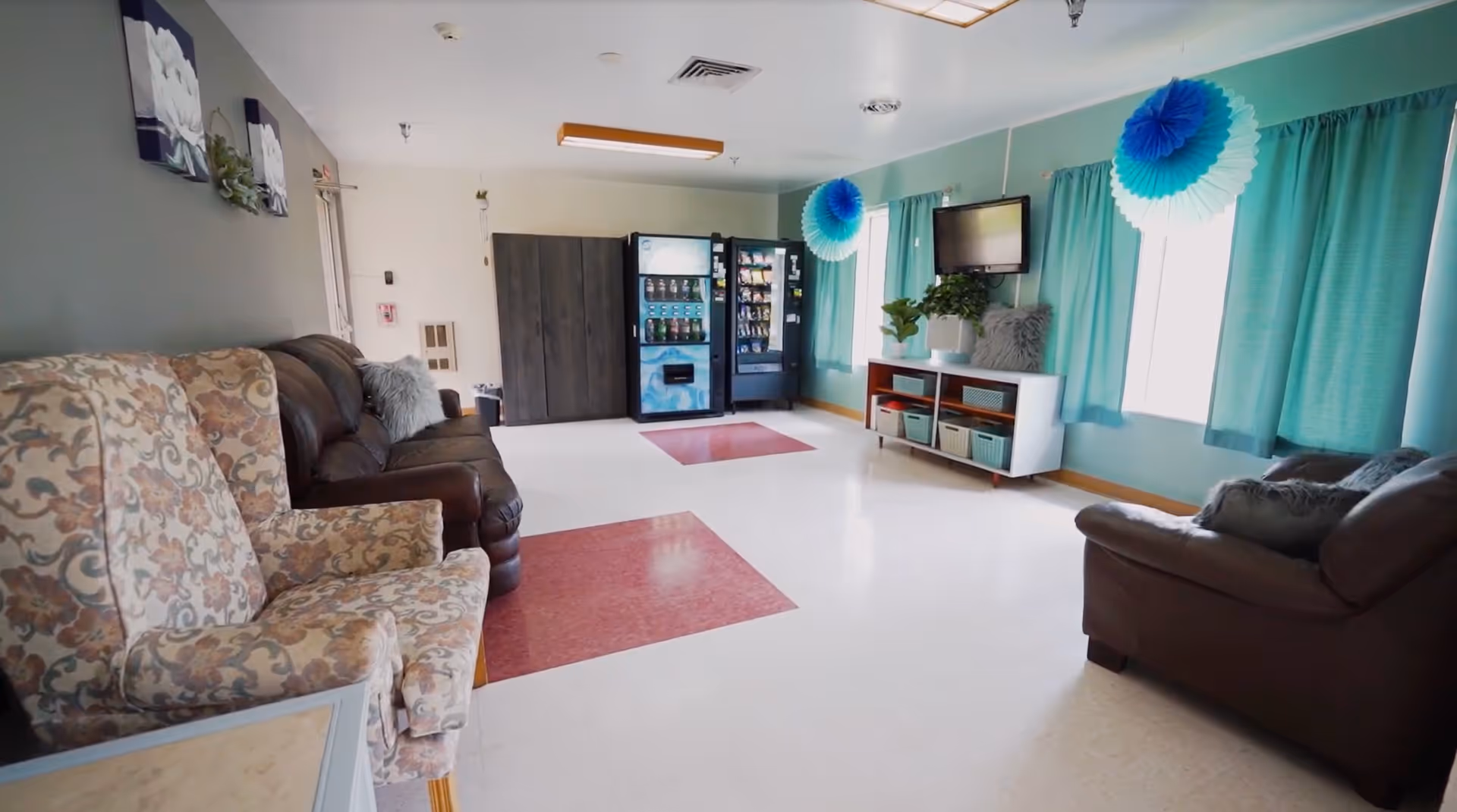 A bright common area with patterned armchairs and brown leather sofas arranged along the walls. The room has teal curtains on the windows, blue and white paper decorations hanging from the ceiling, a small white cabinet with baskets and a potted plant, and a wall-mounted TV. At the far end, there are vending machines and a dark wooden cabinet. The floor is white with red square accents.