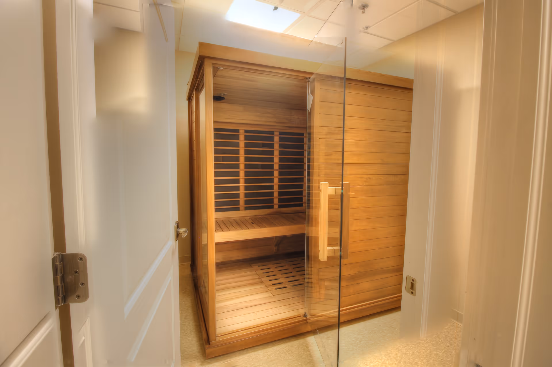 Interior view of a small wooden sauna room with a glass door and wooden benches inside, located in a hallway with white walls and doors.