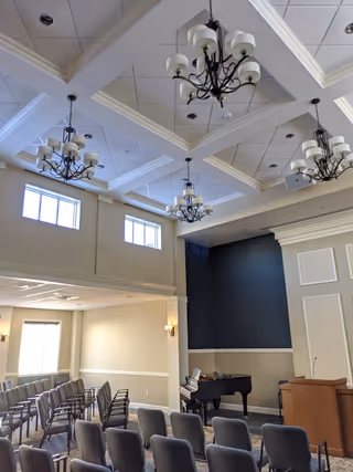 Spacious multipurpose meeting/chapel room with rows of chairs, chandeliers, a piano and a podium beneath a coffered ceiling.