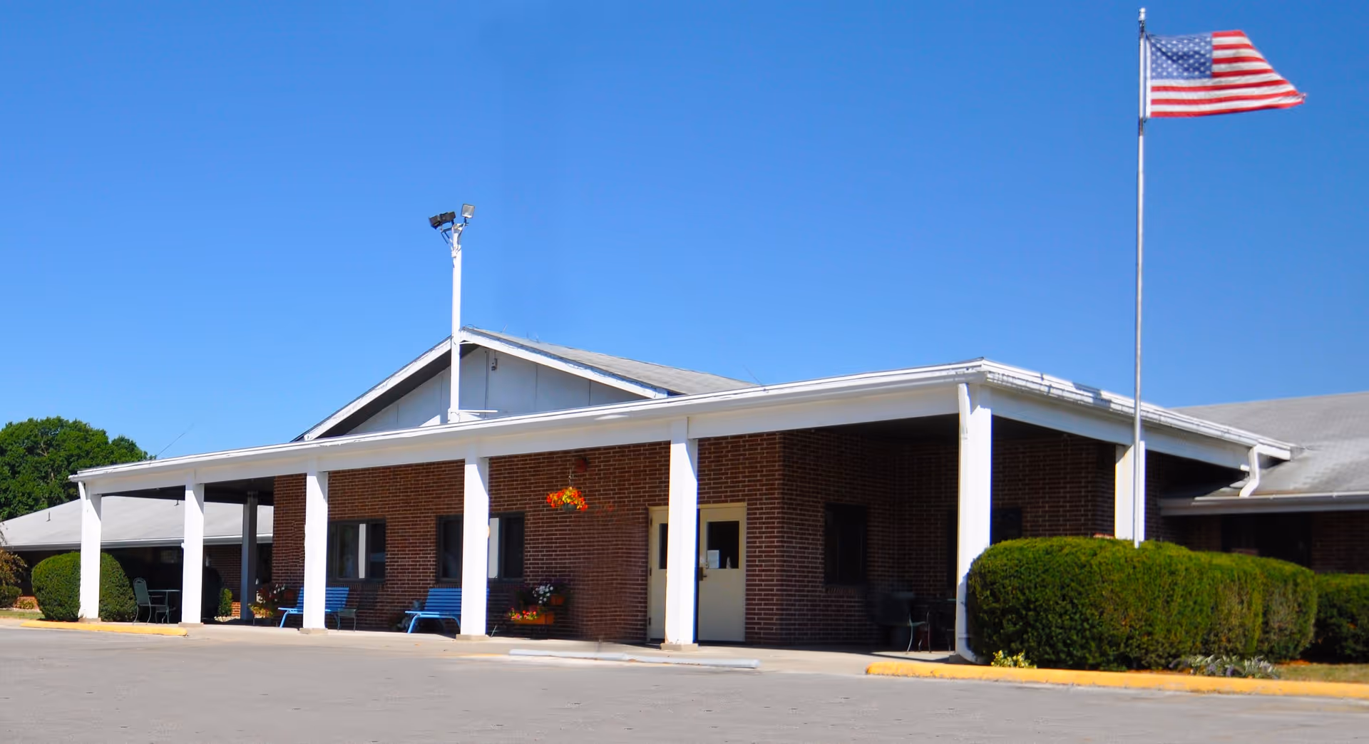 Brick senior living facility exterior with a covered entrance, benches, and an American flag.