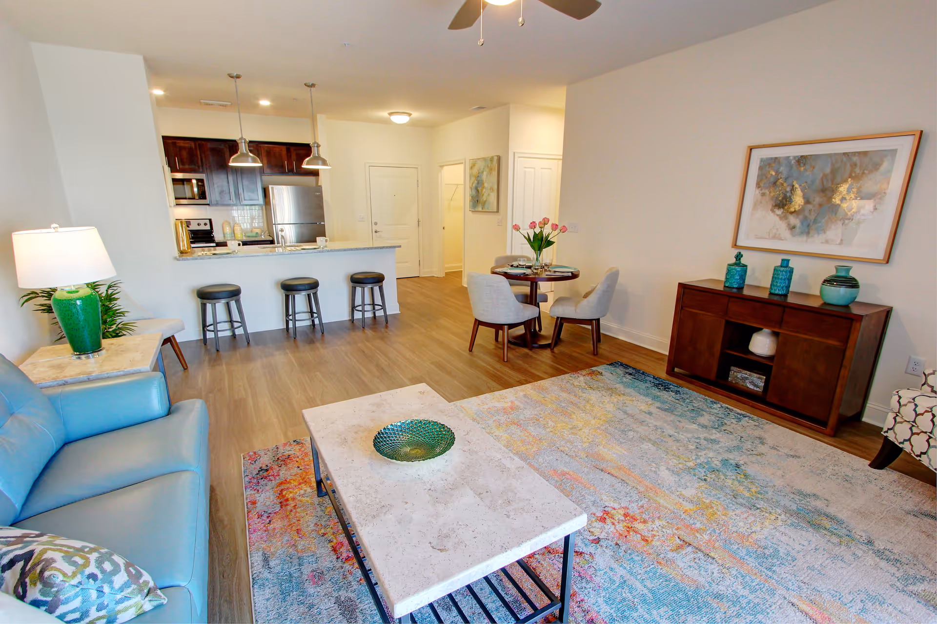 A bright and modern senior apartment living area featuring a light blue sofa with a patterned pillow, a marble coffee table with a decorative green bowl, and a colorful area rug. The room includes a small dining table with four gray upholstered chairs and a vase of pink tulips. The kitchen area has dark wood cabinets, stainless steel appliances, and a granite countertop with three bar stools. The walls are decorated with abstract artwork and the floor is wood laminate.
