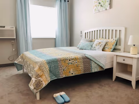A bright bedroom with a white bed frame and a colorful quilt in shades of blue, yellow, and beige. The bed is neatly made with several decorative pillows. Next to the bed is a white nightstand with a small lamp. Light blue curtains frame a window letting in natural light. A pair of blue slippers is placed on the carpeted floor near the foot of the bed.