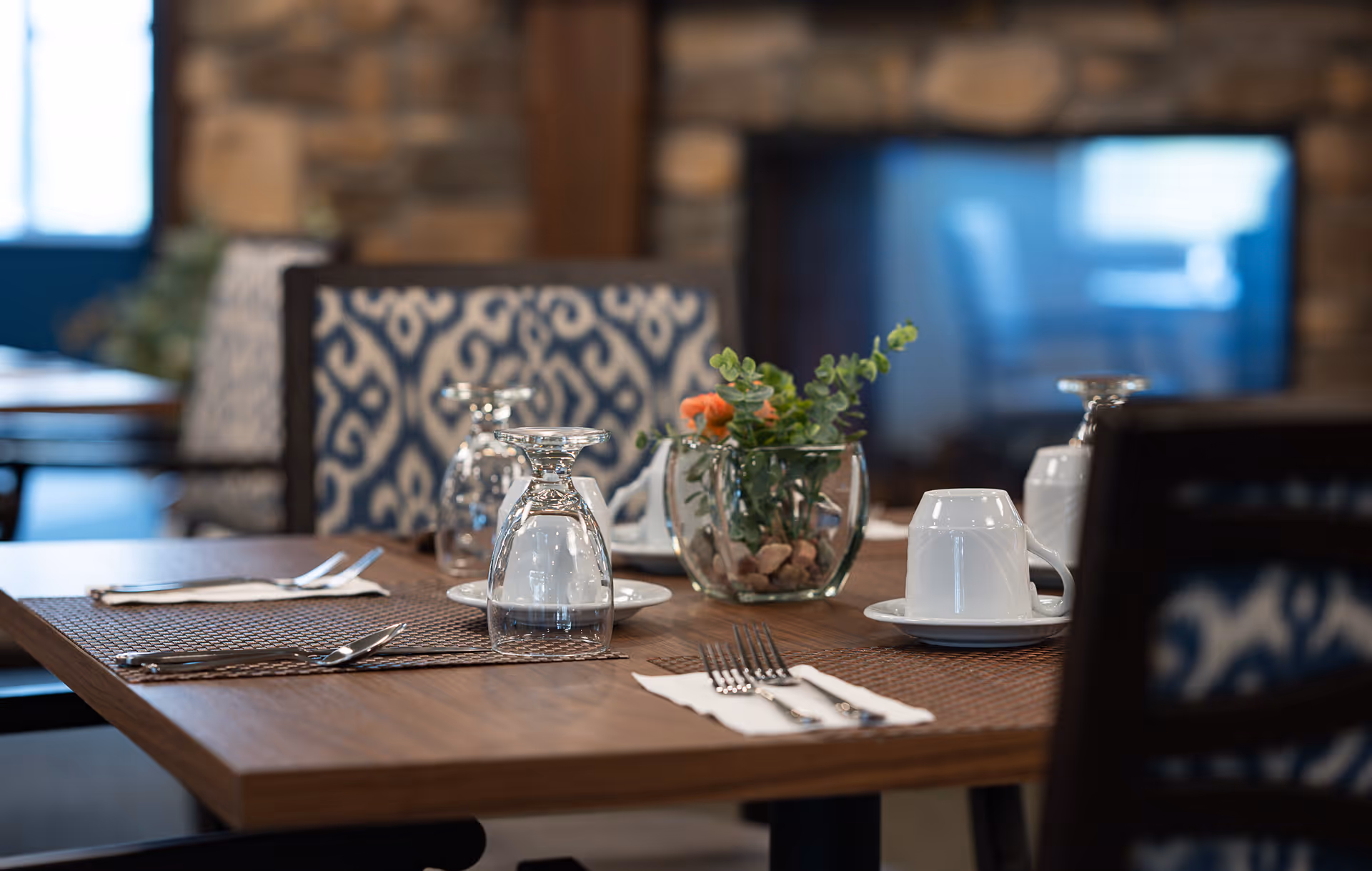 A dining table set with upside-down wine glasses, cups, cutlery and a small floral centerpiece in a cozy dining room.