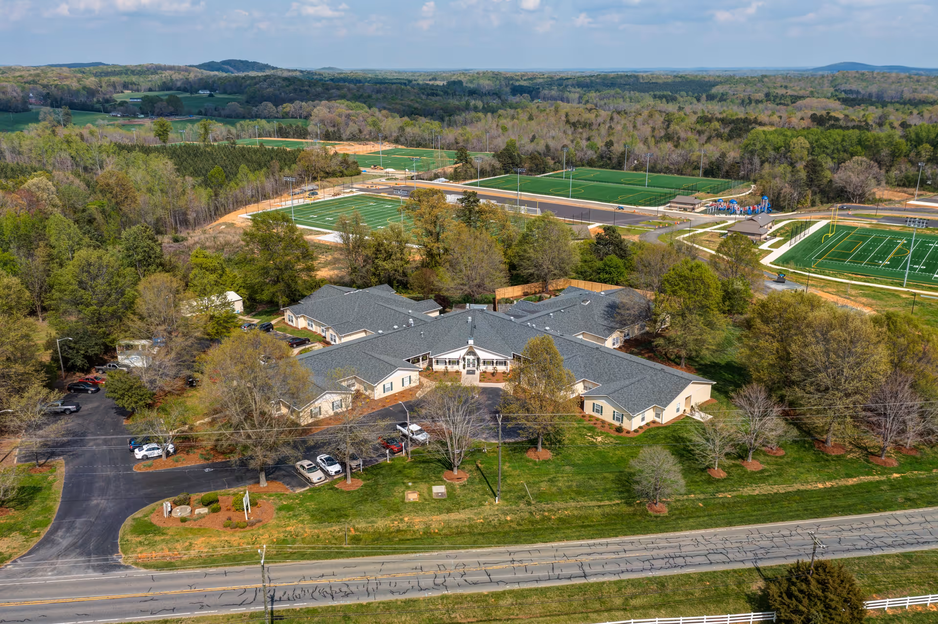 Aerial view of TerraBella Asheboro facility surrounded by trees and greenery, with multiple sports fields including football and soccer fields visible in the background under a partly cloudy sky.