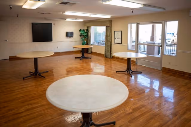 A spacious room with polished wooden floors and four round tables spaced apart. The room has a window with curtains, a door leading outside, a small wall-mounted TV, and a potted plant in the corner.