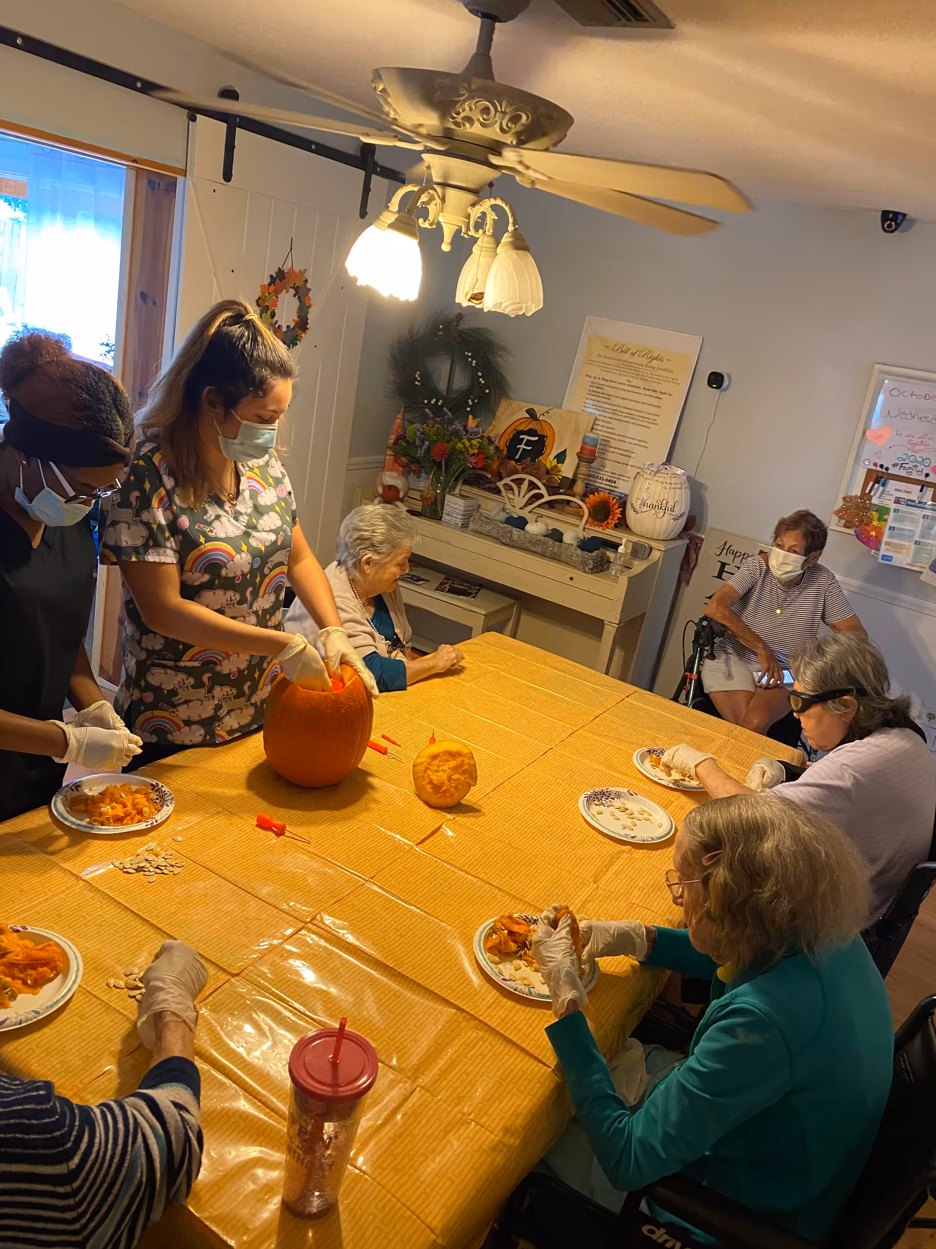 Staff and elderly residents wearing masks and gloves sit around a long table carving pumpkins in a decorated communal dining/activity room.