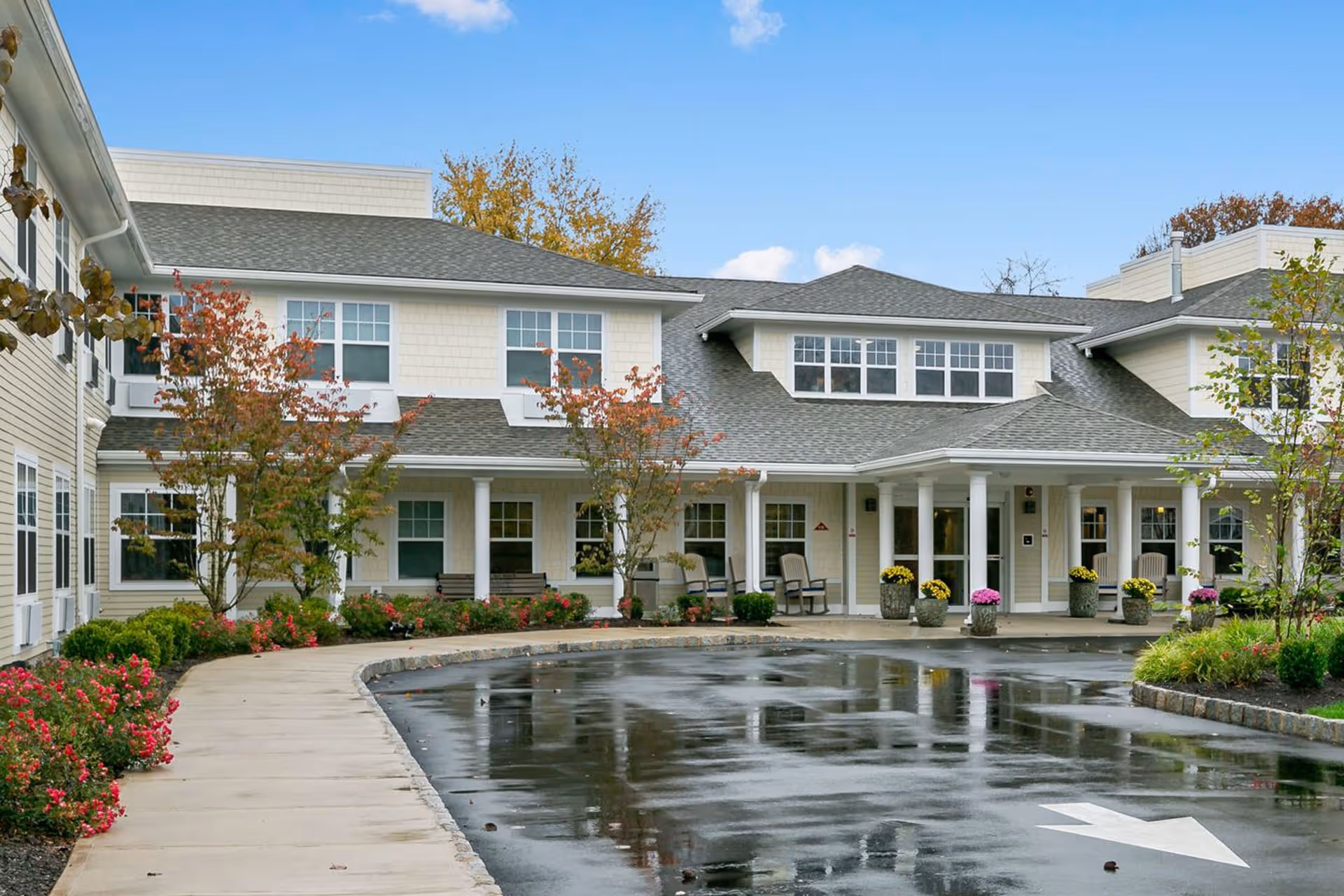 Exterior view of a senior living facility named All American with a paved driveway, landscaped garden beds with flowers and small trees, and a covered entrance supported by white columns. The building has beige siding and multiple windows under a gray shingled roof.