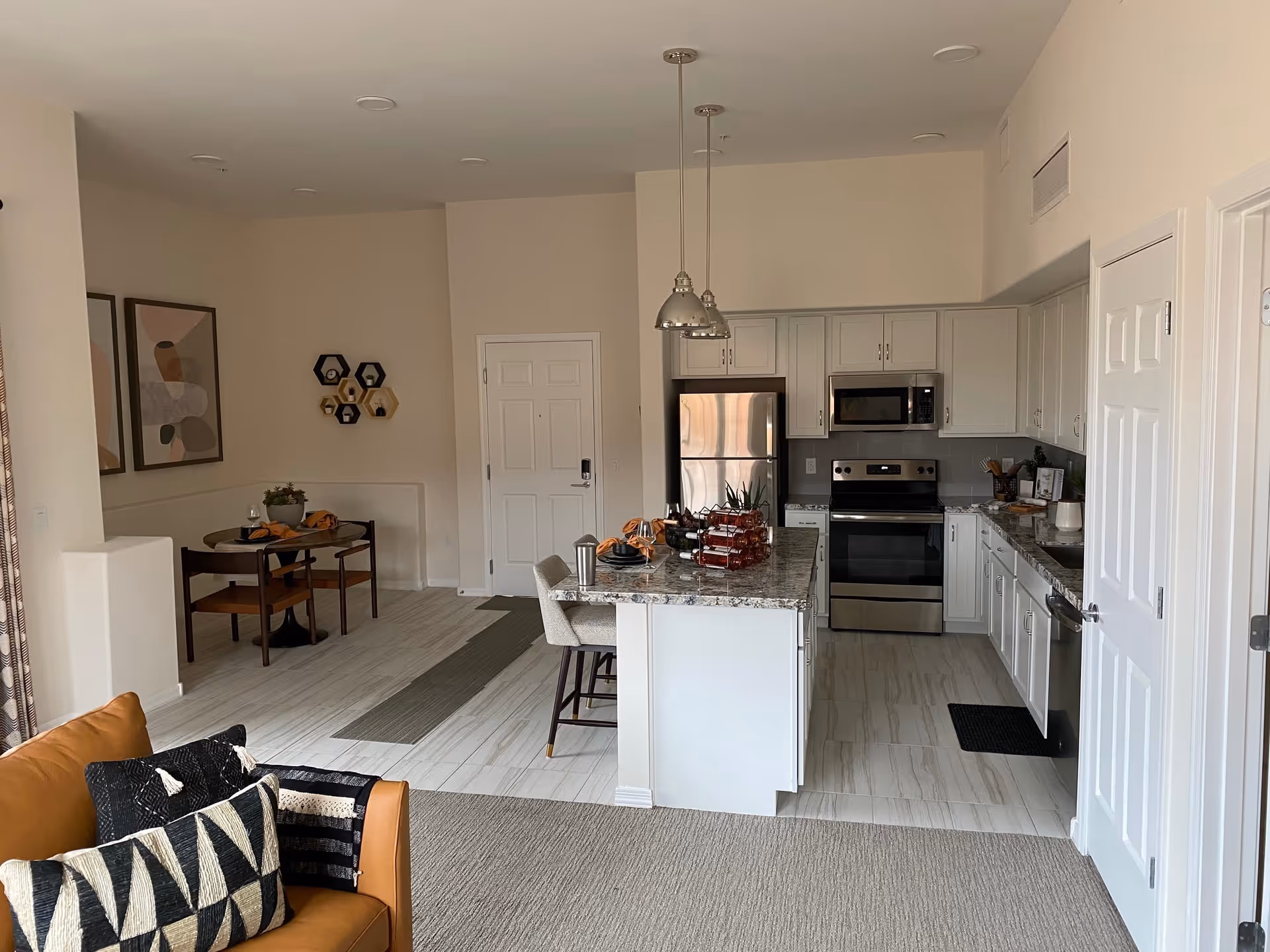 Interior view of a modern kitchen and dining area in a senior living facility. The kitchen features white cabinets, stainless steel appliances including a refrigerator, oven, and microwave, and a granite countertop island with two chairs. The dining area has a small round table with two chairs, decorated with a plant and table settings. A brown sofa with patterned pillows is partially visible in the foreground.