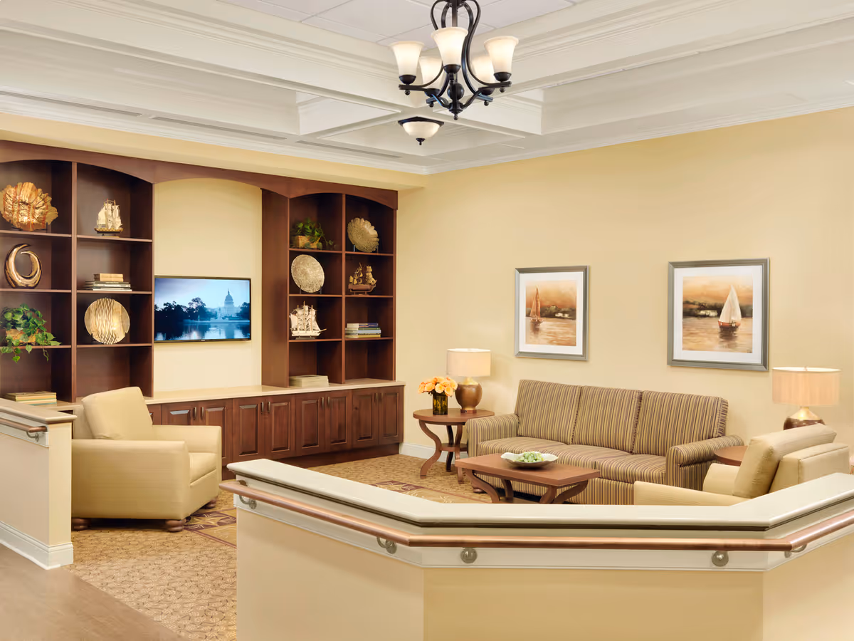 A cozy living room area with beige walls and carpeted floor. The room features a striped sofa, two beige armchairs, a wooden coffee table, and side tables with lamps. A built-in wooden shelving unit with decorative items and a flat-screen TV is mounted on the wall. Two framed paintings of sailboats hang on the wall above the sofa. A chandelier with multiple lights hangs from the ceiling.