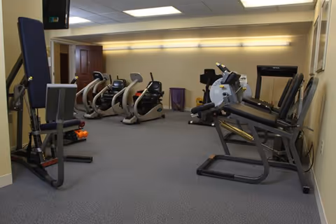 A small indoor exercise room with various fitness equipment including stationary bikes, a leg press machine, and other workout machines. The room has beige walls, a gray carpeted floor, and fluorescent lighting on the ceiling.