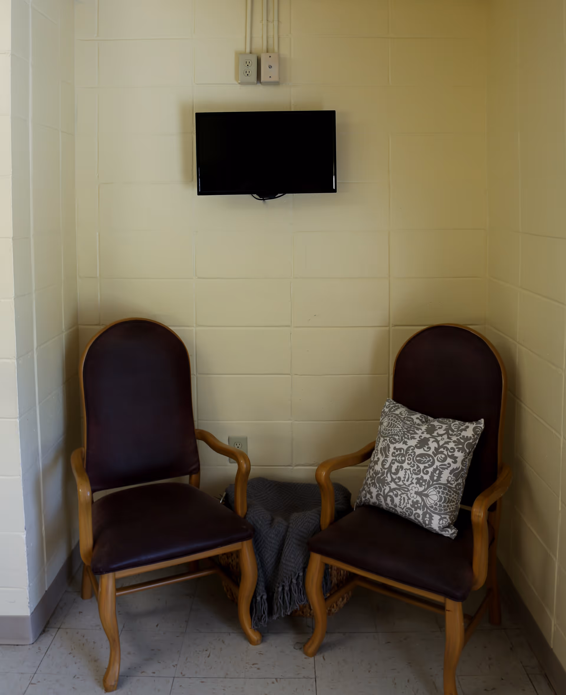 Two wooden armchairs with dark cushions are placed against a beige cinder block wall. One chair has a decorative patterned pillow. Between the chairs is a basket with a folded gray blanket. A small flat-screen TV is mounted on the wall above the chairs.