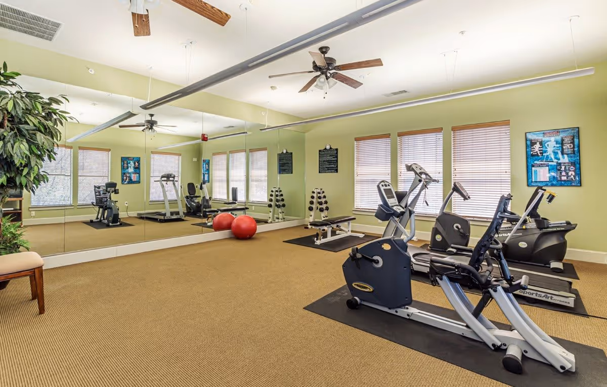 A fitness room with exercise equipment including stationary bikes, a treadmill, a weight bench, dumbbells, and two red exercise balls. The room has large mirrors on one wall, several windows with blinds, green walls, and ceiling fans.