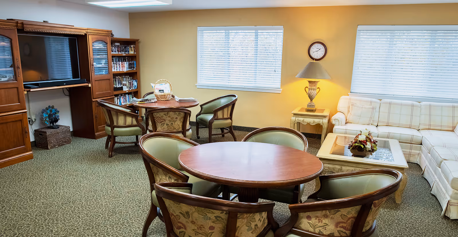 A cozy senior living community common room with two round wooden tables surrounded by upholstered chairs, a large wooden entertainment center with a TV and shelves filled with DVDs, a plaid sectional sofa, a glass-top coffee table with a floral arrangement, a side table with a lamp, and two windows with blinds letting in natural light.