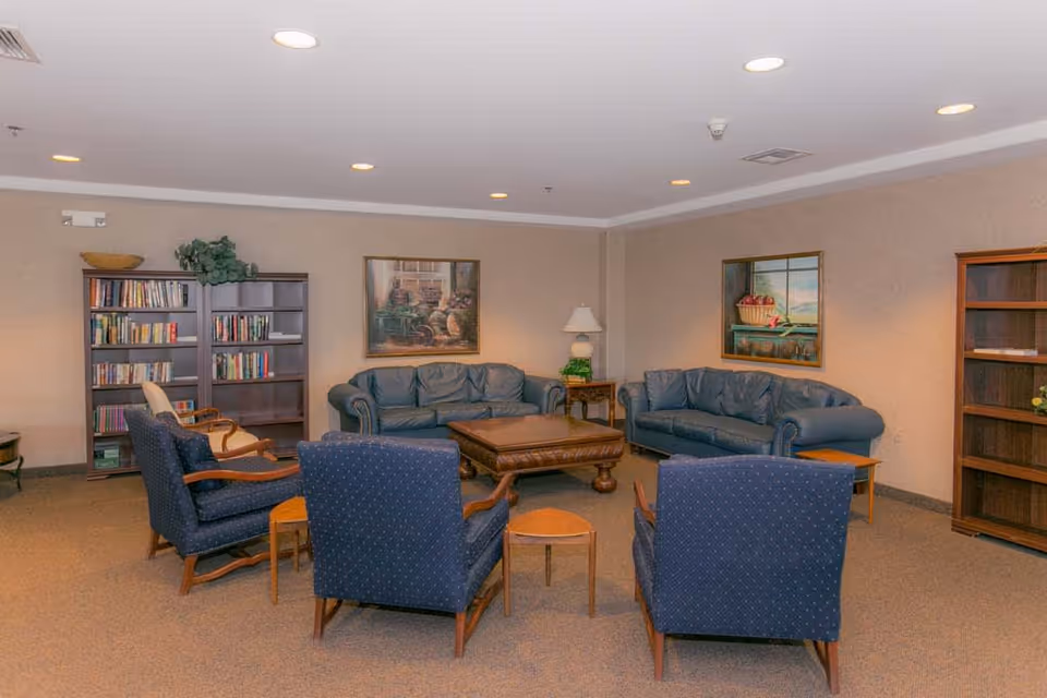 A cozy living room in Mountain View Retirement Village featuring two blue leather sofas, four blue upholstered armchairs arranged around a wooden coffee table, two wooden bookshelves filled with books, a side table with a lamp, and framed paintings on the beige walls.