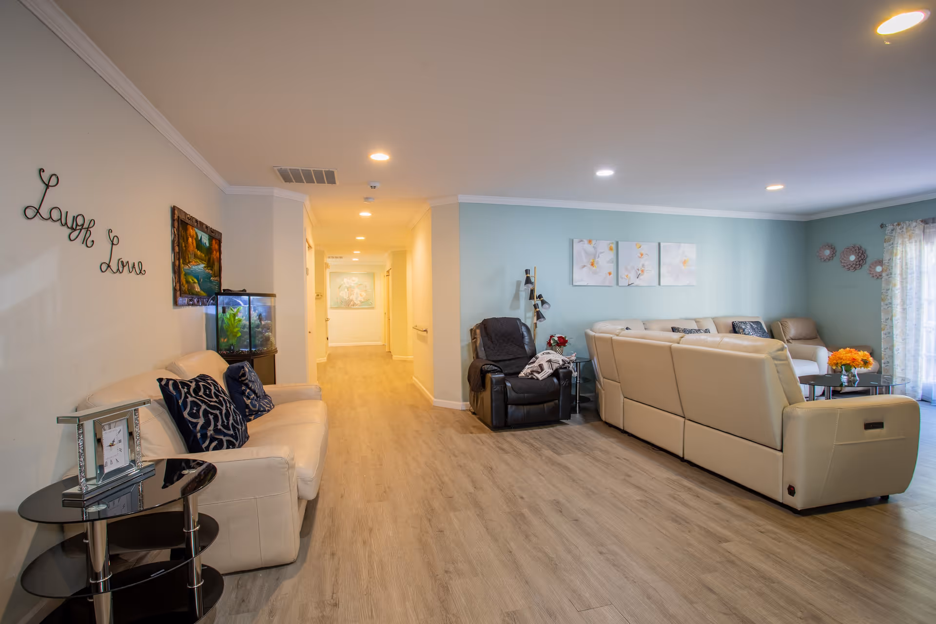 A spacious living room area with light wood flooring, beige leather sofas, a black recliner chair, and a small glass side table with a clock. The walls are painted light blue and white, decorated with floral artwork and metal wall art. There is a fish tank on a stand next to a beige loveseat with dark blue patterned pillows. The room is well-lit with recessed ceiling lights and natural light coming through a window with sheer curtains.
