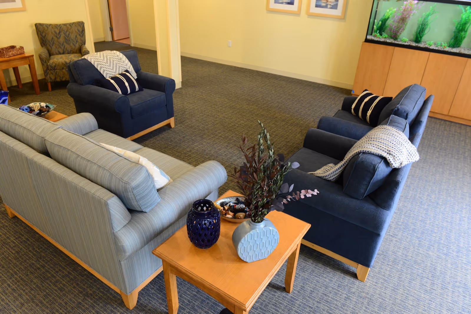 A cozy living room area with two blue armchairs, a light blue striped sofa, and a wooden coffee table with decorative items including a blue vase and a plant. There is a large fish tank on a wooden cabinet against the wall and framed pictures hanging above it. The room has carpeted flooring and light yellow walls.