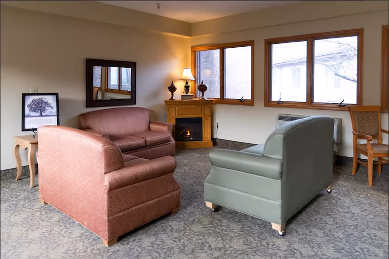 A cozy sitting area in a retirement community with two upholstered sofas facing each other, a wooden side table with a framed picture of a tree, a wall mirror, a corner fireplace with a lit lamp and decorative items on the mantel, and three large windows letting in natural light.