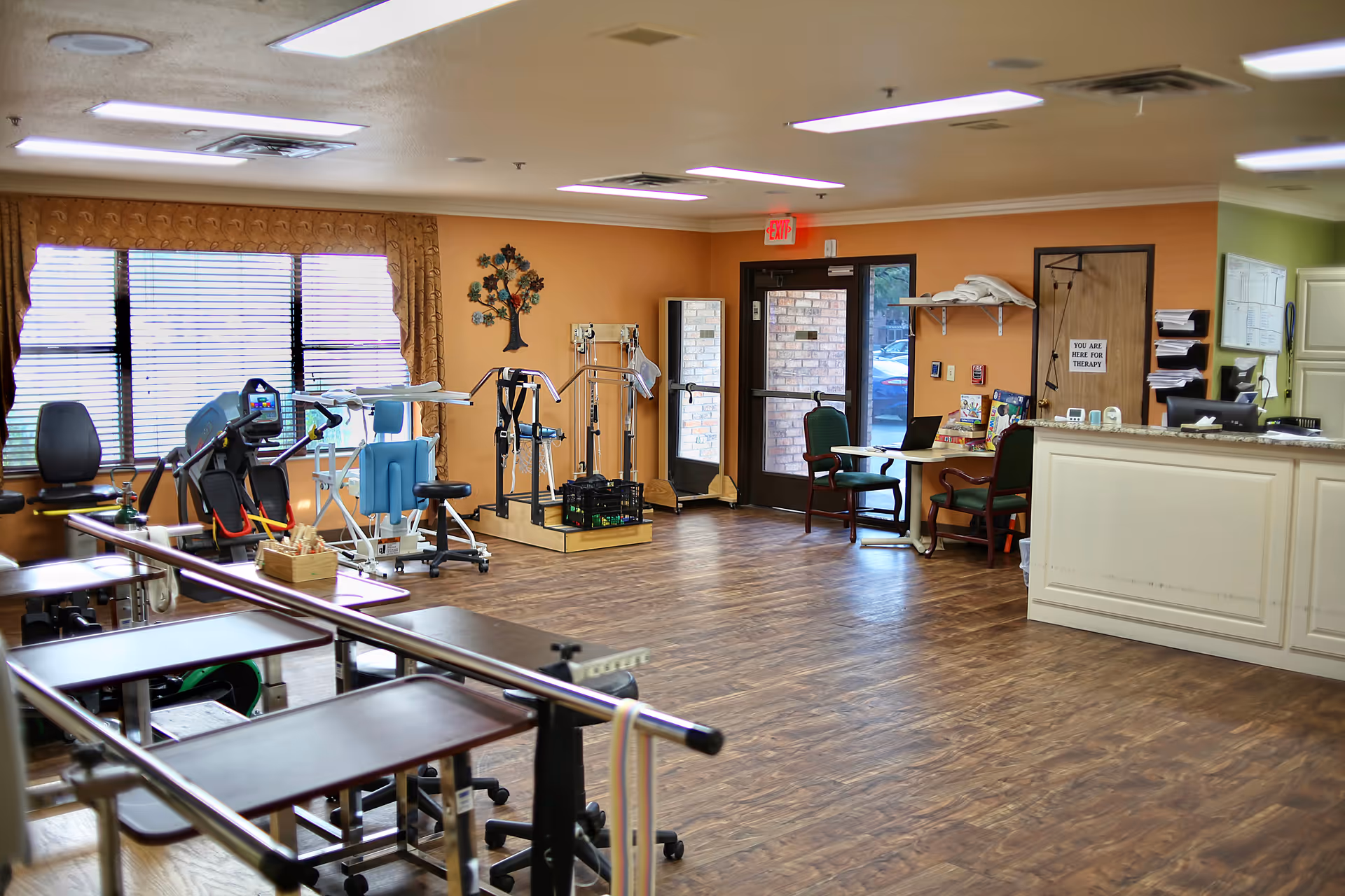 Interior view of a therapy room in a nursing center with exercise equipment, parallel bars for walking practice, chairs, a reception desk, and a door leading outside. The room has wood flooring, orange and green walls, and large windows with blinds.