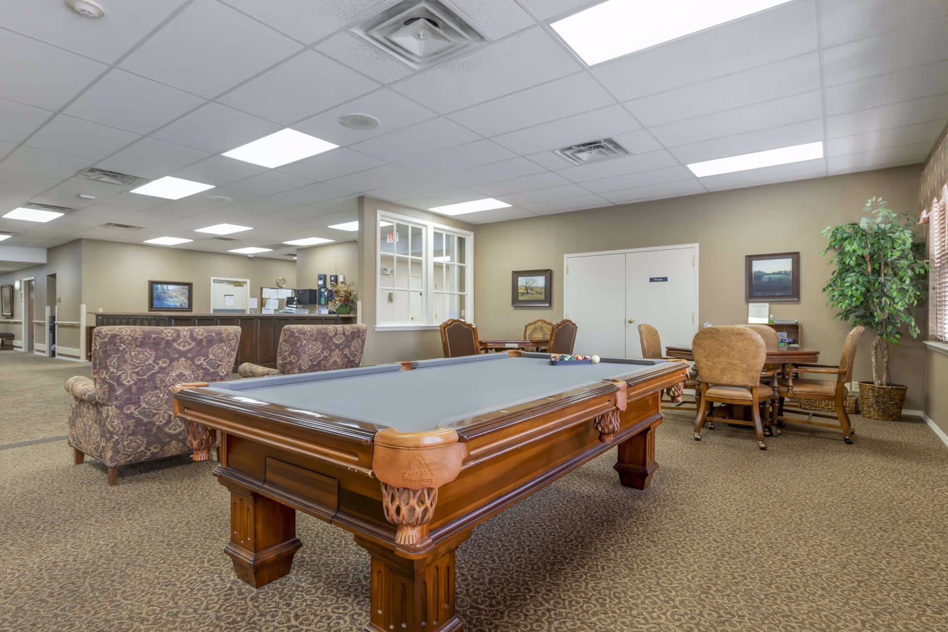 A spacious common area in Aberdeen Heights Assisted Living featuring a pool table with billiard balls and cue ball on it. The room has patterned carpet, beige walls, and a drop ceiling with fluorescent lights. There are upholstered armchairs, a table with chairs, framed paintings on the walls, a potted plant near the window, and a reception desk in the background.