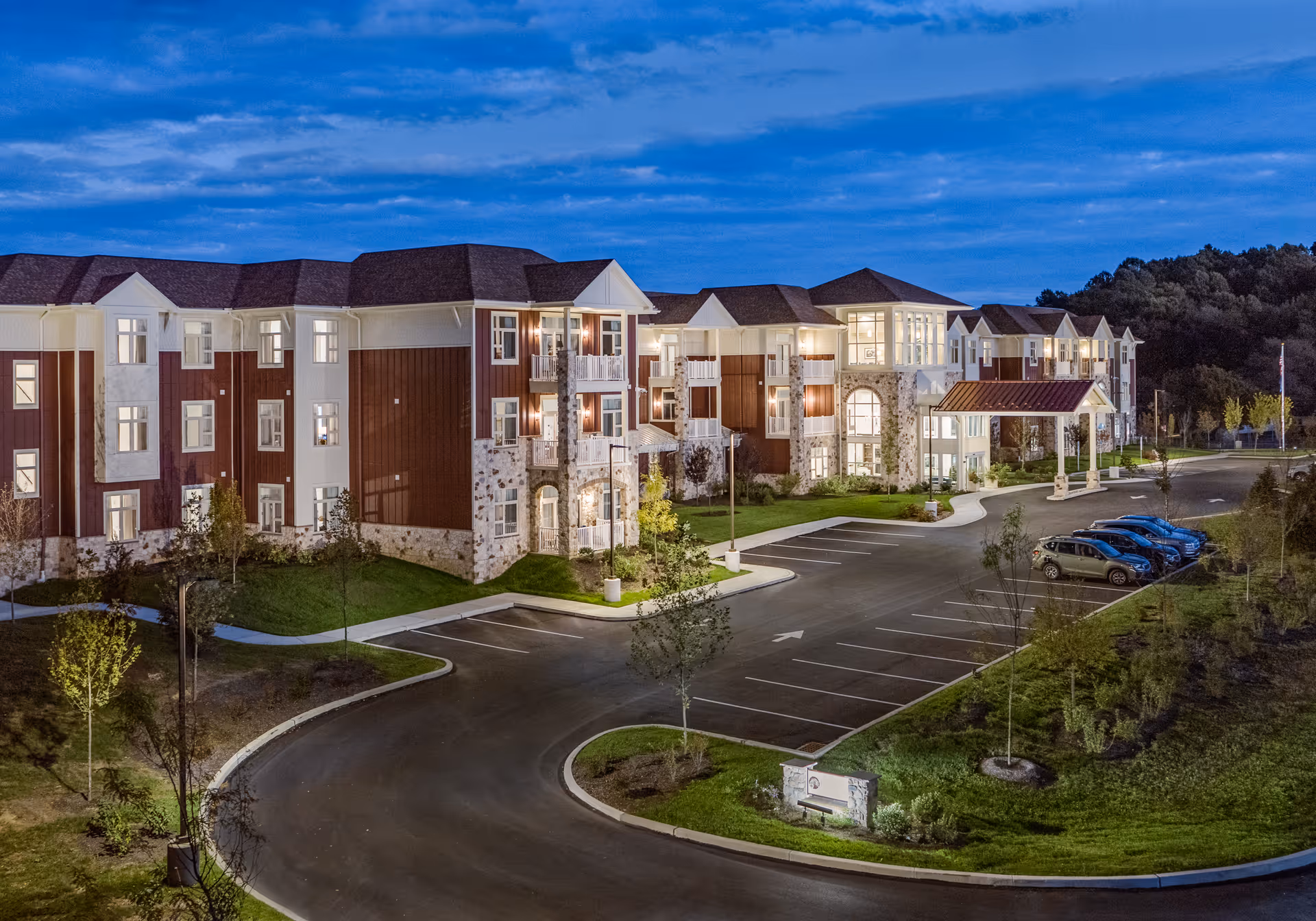 Exterior view of Fieldstone at Chester Springs senior living facility at dusk, showing a large three-story building with illuminated windows, a covered entrance, a curved driveway, parking spaces with several cars, and landscaped greenery around the property.