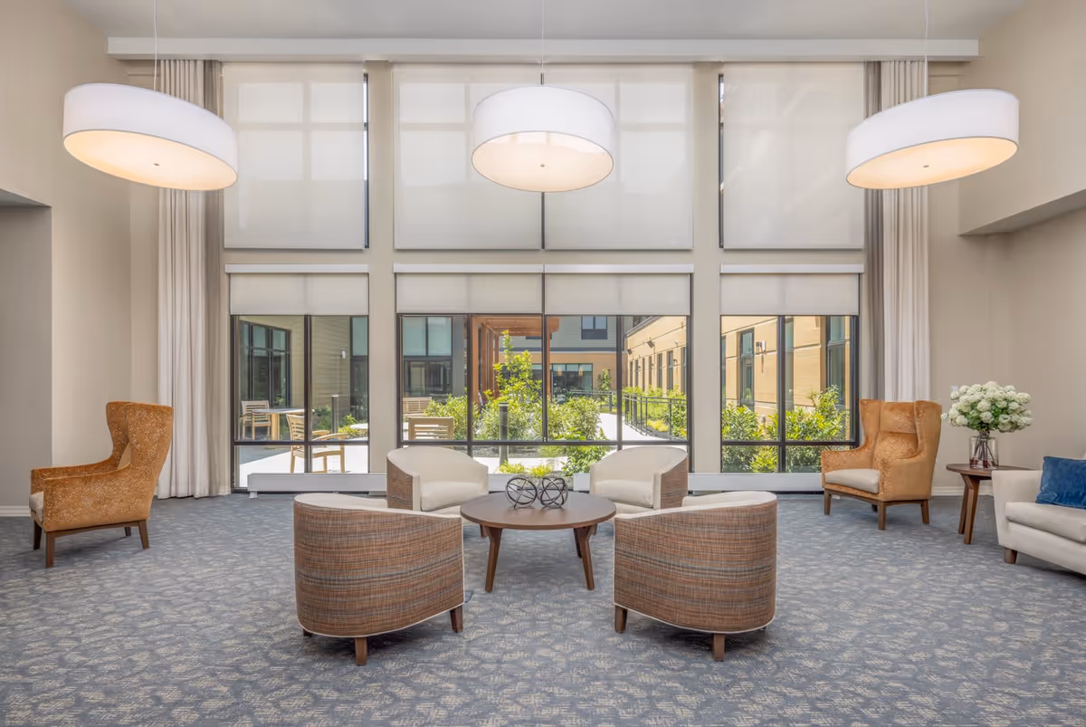 A bright and spacious sitting area with large windows overlooking a garden courtyard. The room features four cushioned chairs arranged around a round wooden coffee table with decorative objects on it. Two orange wingback chairs are positioned on either side near the windows, and a beige sofa with a blue pillow is partially visible on the right. Three large pendant lights hang from the ceiling, and the floor is carpeted with a patterned design.