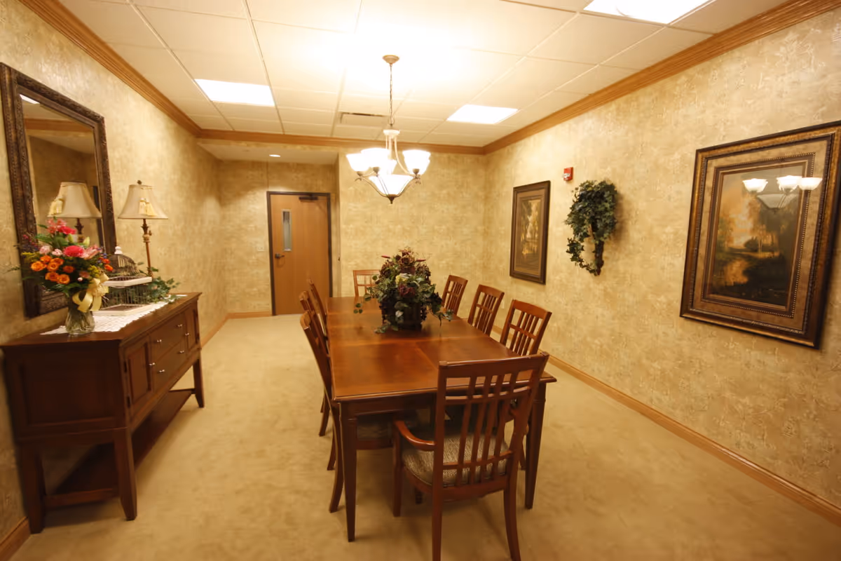 A formal dining room with a long wooden table surrounded by eight wooden chairs. The table has a floral centerpiece. The room has beige patterned wallpaper, a beige carpet, and a wooden sideboard with a lamp, flowers, and decorative items. Two framed paintings and a wreath hang on the walls. A chandelier with multiple lights hangs above the table, and a wooden door is visible at the far end of the room.