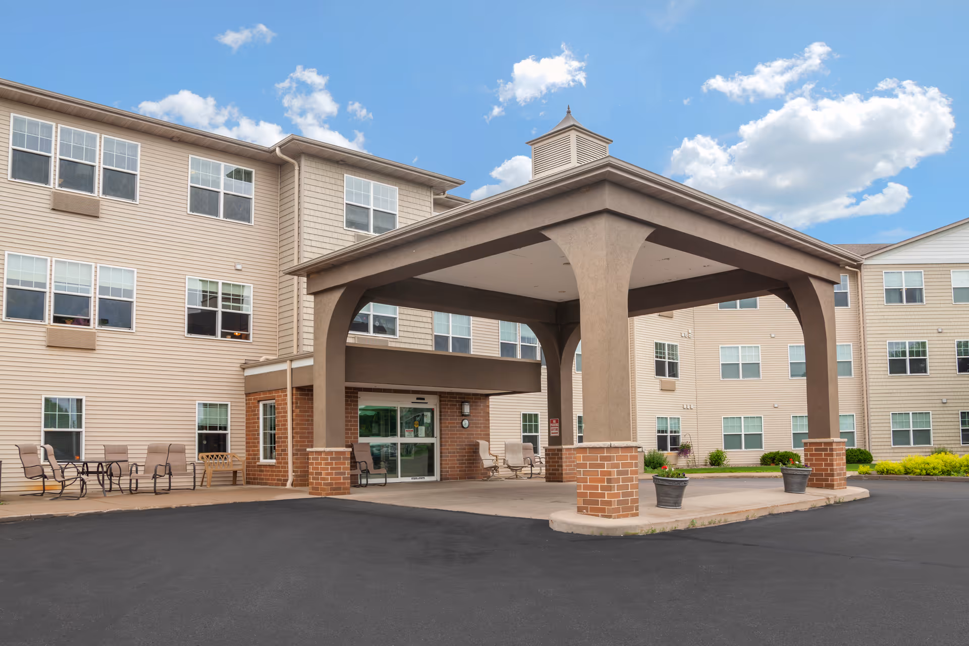 Front entrance of a beige multi-story senior living building with a covered porte-cochere, outdoor seating, and planters.