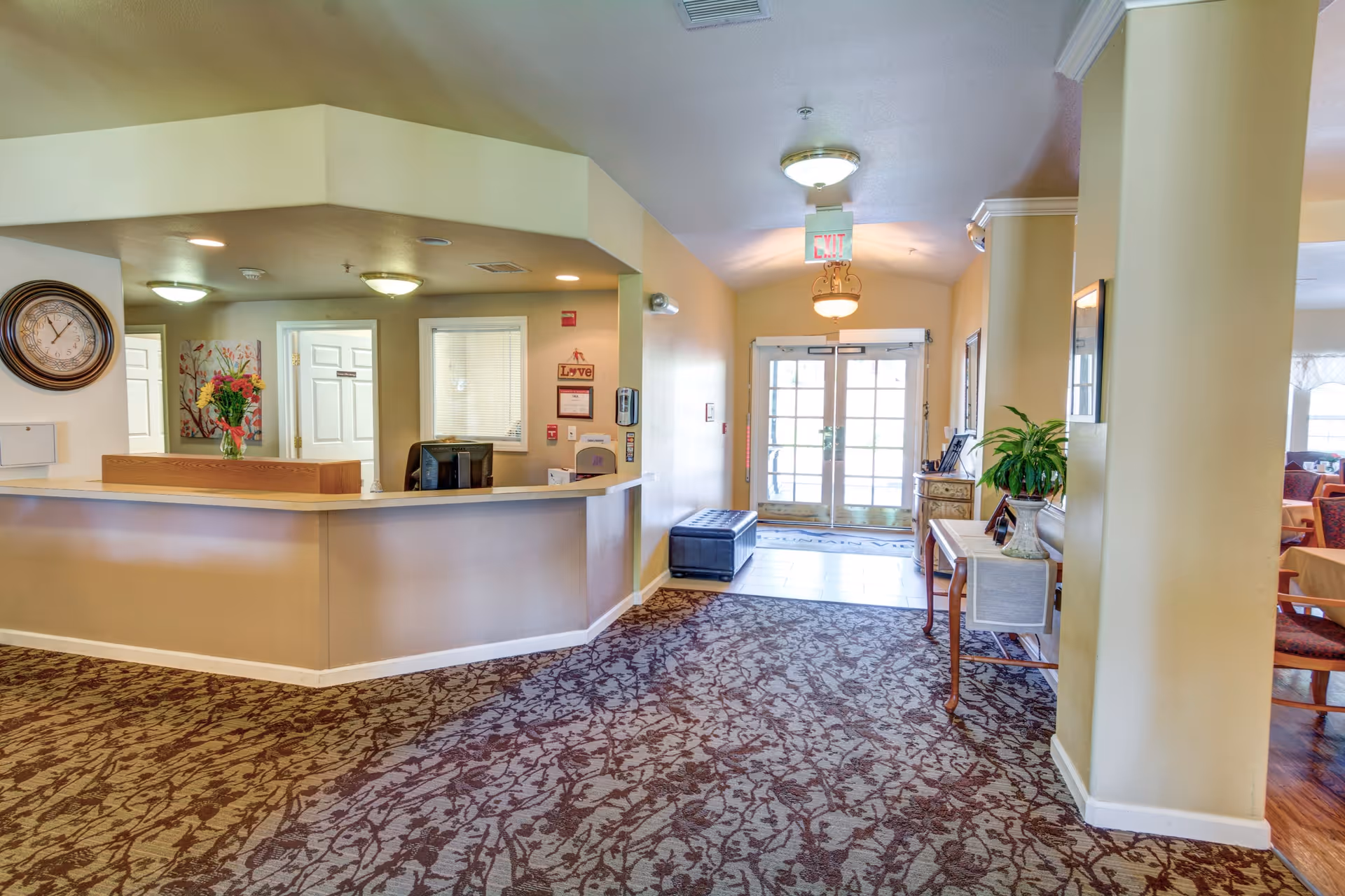 Interior view of a senior living facility reception area with a curved front desk, a wall clock, floral artwork, and a vase with flowers on the counter. The area has patterned carpet flooring, beige walls, and ceiling lights. There is a hallway leading to double glass doors with an exit sign above them. To the right, there is a small table with a plant and framed pictures, and a glimpse of a dining area with tables and chairs.